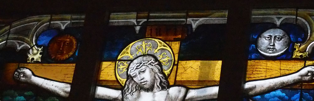 Partie haute d'un vitrail avec une réprésentation de la Crucifixion. Jésus est sur la croix et au-dessus de lui, à gauche et à droite sont respectivement deux cercles dans lesquels des visages dorés et gris évoquant le soleil et la lune.